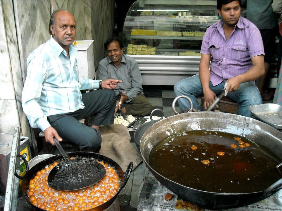 Gulab jamuns frying slowly in a wide pan of oil next to a pot of sugar syrup at a sweet shop
